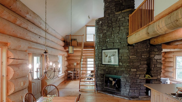 Interior de una cabaña de troncos con comedor, chimenea de piedra, escalera de madera y un dormitorio en lo alto.