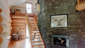 Interior de una cabaña de troncos con una escalera de madera, una chimenea de piedra y un cuadro de un ciervo enmarcado.