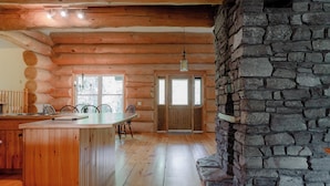 Interior de una cabaña de troncos con chimenea de piedra, comedor de madera y cocina con isla central.