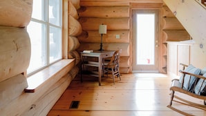 Interior de una cabaña de troncos con una mesa y sillas de madera, una lámpara y una ventana con vistas.