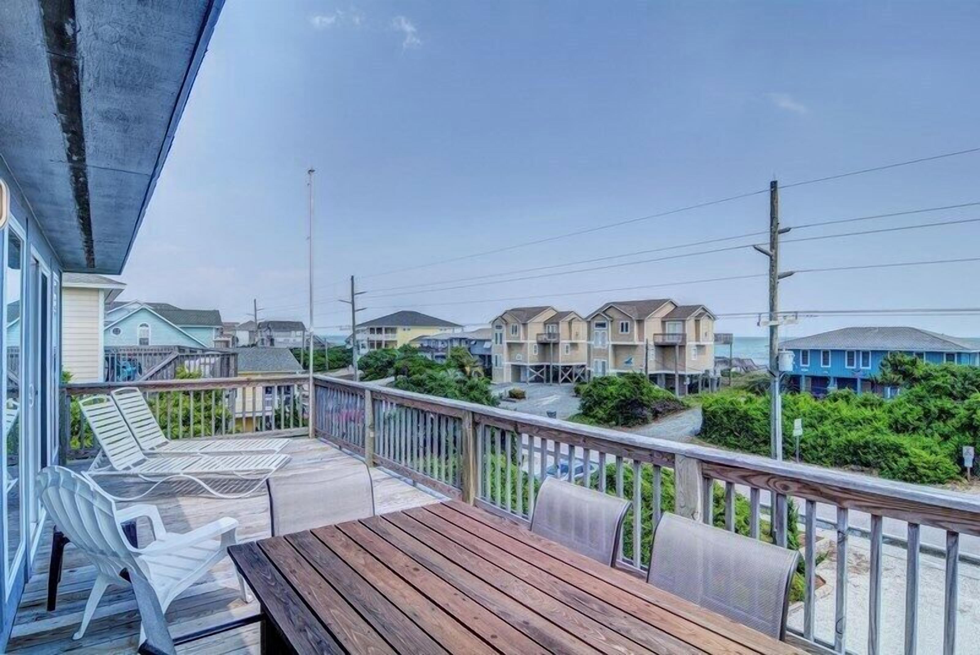 A wooden deck with a table and chairs, overlooking a coastal residential area with houses and a clear sky.