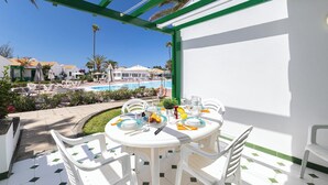 A sunlit outdoor dining area with a pool view, white furniture, and a green-framed pergola.
