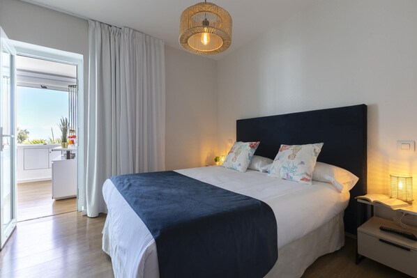 A bedroom with a large bed, a bedside table with a lamp, a window with curtains, and a view of the outside.