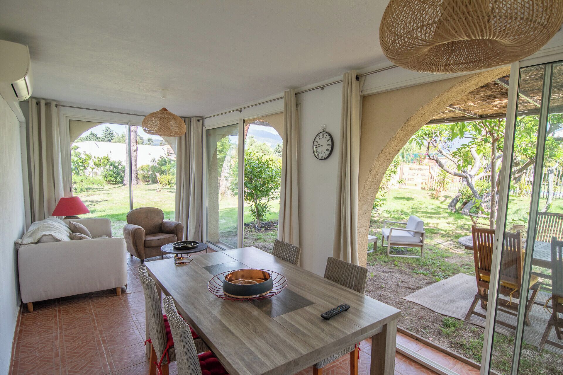 Une pièce spacieuse comprenant une grande table à manger, un canapé et un fauteuil. Un réveil est accroché au mur et une fenêtre offre une vue sur l’extérieur.