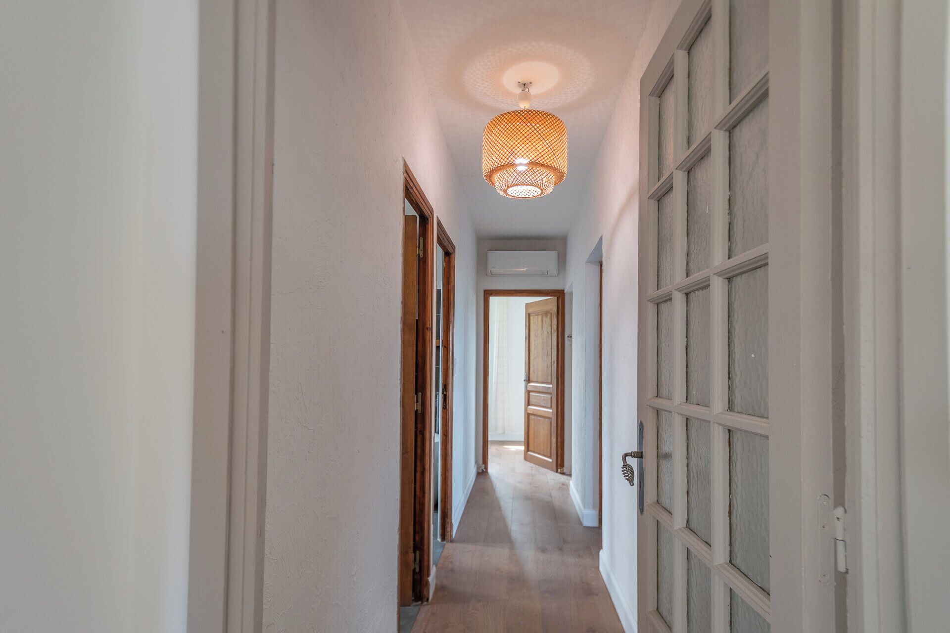 Un pasillo con una puerta de madera a la derecha, un lámpara colgante y un techo blanco.
