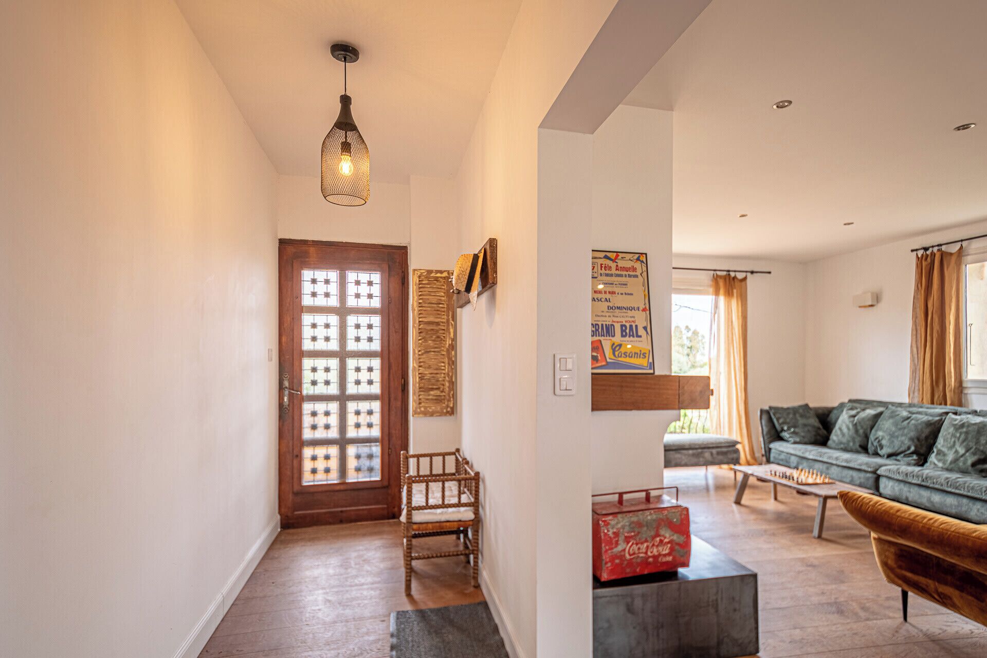 Un salon avec une porte en bois, un fauteuil en osier, un canapé, une table basse et un luminaire suspendu.