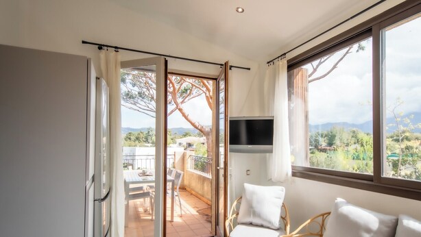 Een kamer met een schuifglazen deur naar een terras buiten, een wandgemonteerde flatscreen-tv en twee witte fauteuils.