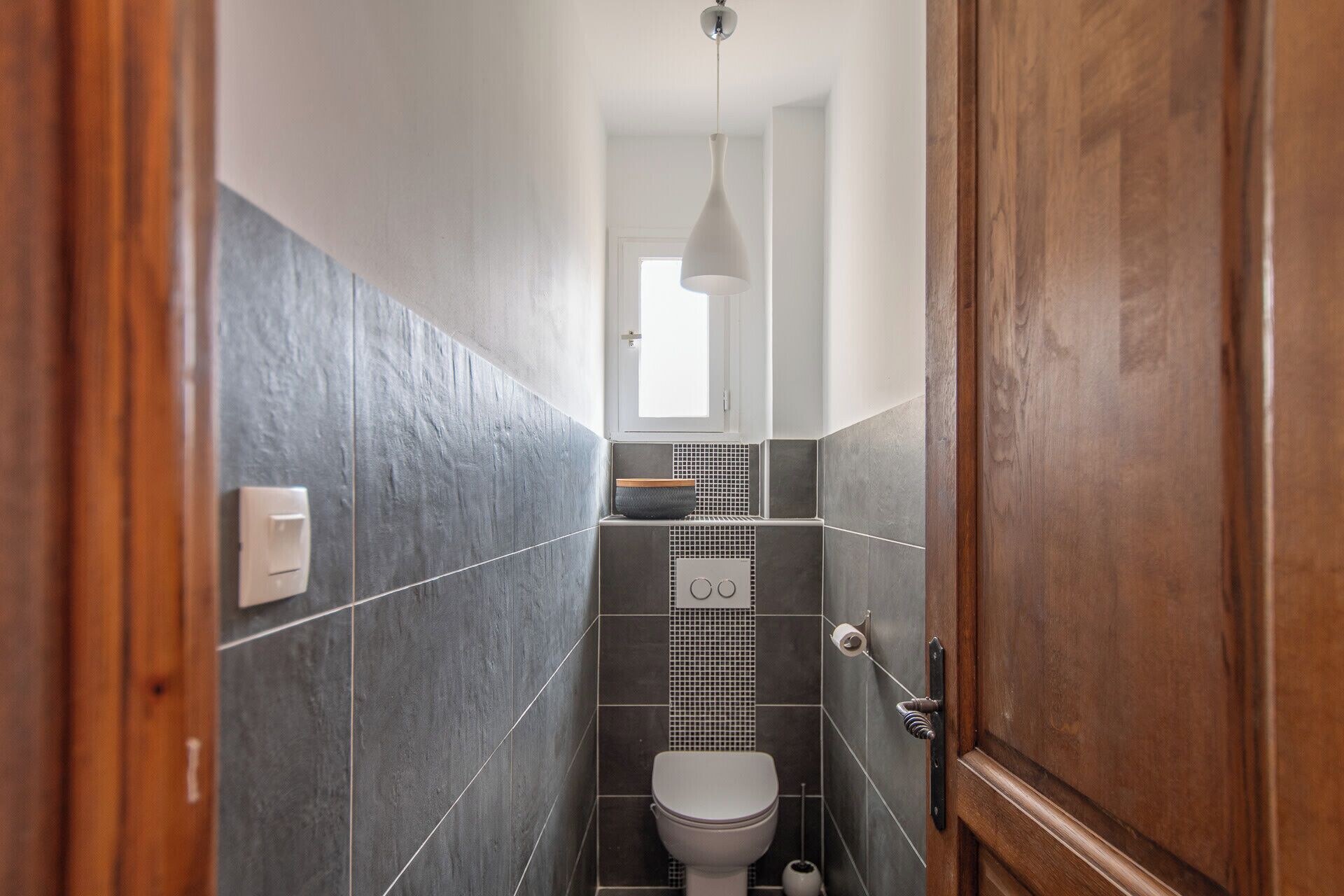 Un pasillo estrecho que conduce a un baño con inodoro, un lavamanos pequeño y un salpicadero de azulejos.