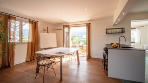 Ruime kamer met een eettafel, stoelen, een schilderij en een keukenhoek met een spoelbak en barstoelen.