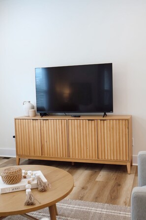 A modern living room with a flat-screen TV on a wooden cabinet, a round coffee table with decorative items, and a light-colored rug.