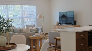 A modern living room with a sofa, dining table, and chairs, a kitchen area with a TV, and a large window with blinds. - Townhouse on Shipley (Cookeville)