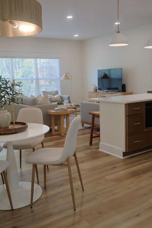 A modern living room with a sofa, dining table, and chairs, a kitchen area with a TV, and a large window with blinds.