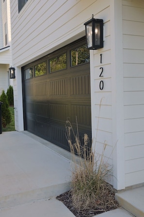 A residential garage with a dark door, a number '120' on the wall, and two wall-mounted lanterns.