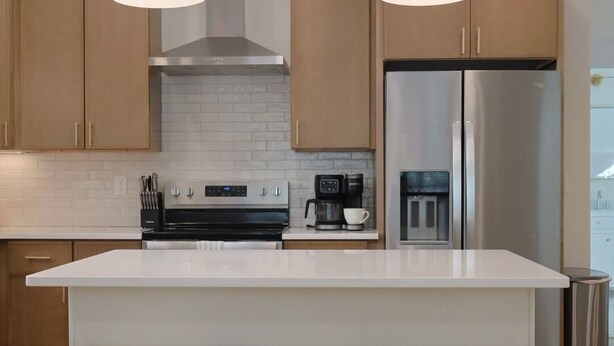 A modern kitchen with a large island, wooden stools, pendant lights, stainless steel appliances, and a white backsplash. - Townhouse on Shipley (Cookeville)