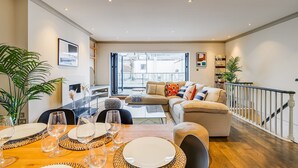 Comedor con mesa de madera dispuesta para cuatro personas, sofá y escalera que sube a un nivel superior.