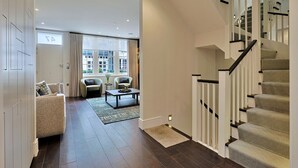 Een ruime woonkamer met een bank, salontafel en uitzicht door een groot raam. Een trap met een patroonbelegd tapijt leidt naar een verdieping.