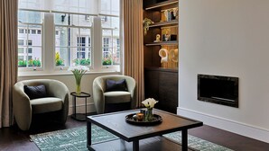Een moderne woonkamer met twee fauteuils, een salontafel, een open haard en uitzicht op gebouwen door grote ramen.
