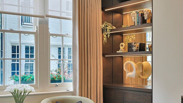 Een moderne woonkamer met een groot raam, een comfortabele fauteuil, een bijzettafeltje met decoratieve items, en een boekenkast vol diverse objecten.