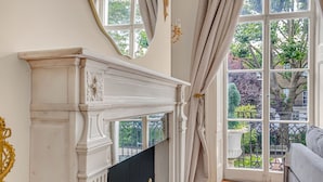Egy fehér kandallóval, nagy, függönnyel ellátott ablakokkal rendelkező szoba, ahonnan a kertre nyílik kilátás.