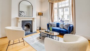 A modern living room with a fireplace, a blue sofa, white armchairs, and a glass coffee table. - Belgravia - Charming Spacious 4 Bed Home for 9 (London)