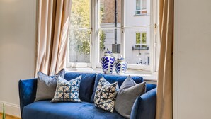 A blue sofa with patterned cushions, a window with curtains, and decorative vases on the windowsill. - Belgravia - Charming Spacious 4 Bed Home for 9 (London)