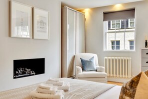 Une chambre à coucher comprenant un lit, une chaise, un radiateur et une fenêtre donnant sur un immeuble.