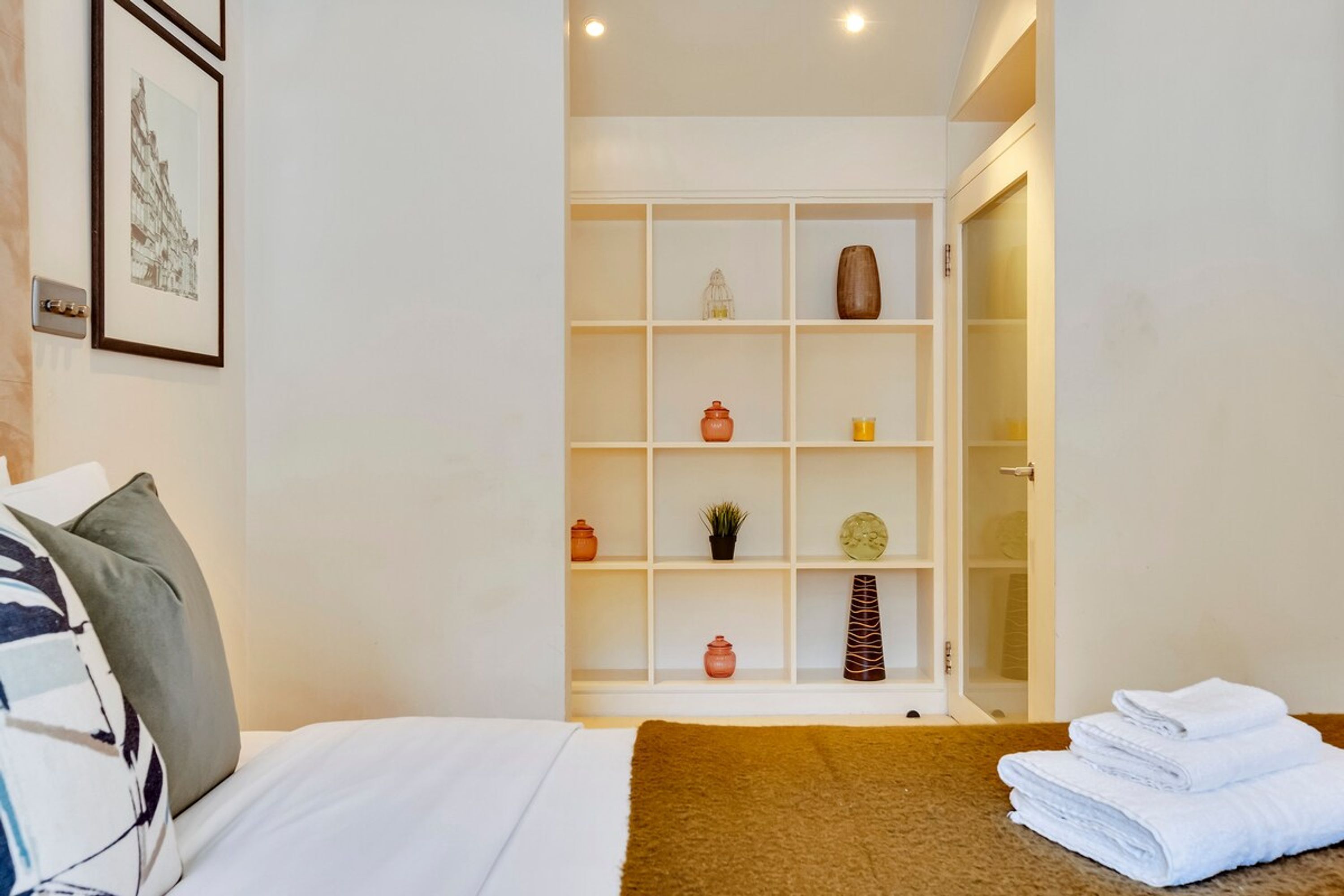 A hotel room with a neatly made bed, a brown throw blanket, and a stack of white towels on the side. There is a white shelving unit with various decorative items and a framed picture on the wall.