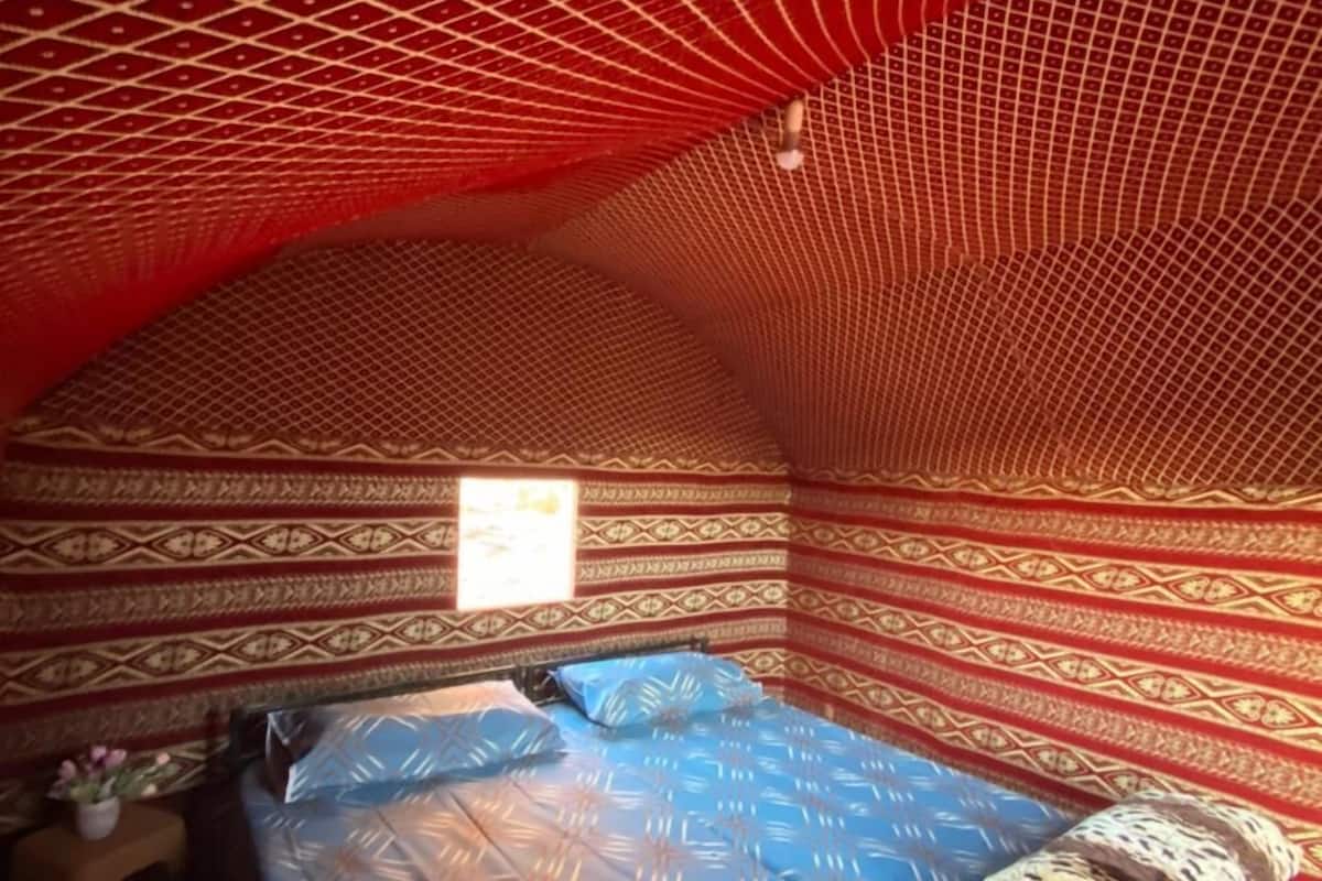 Une pièce avec un mur aux motifs rouges et beiges, un lit recouvert d’une courtepointe bleue à motifs et une petite fenêtre.