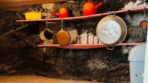 A rustic kitchen with a tiled floor, wooden shelves holding cooking pots, and a hanging light bulb.
