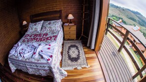 A bedroom with a wooden bed, a nightstand, a lamp, a rug, and a balcony with a view of hills. - Cabanas Mountain (Campos do Jordão)