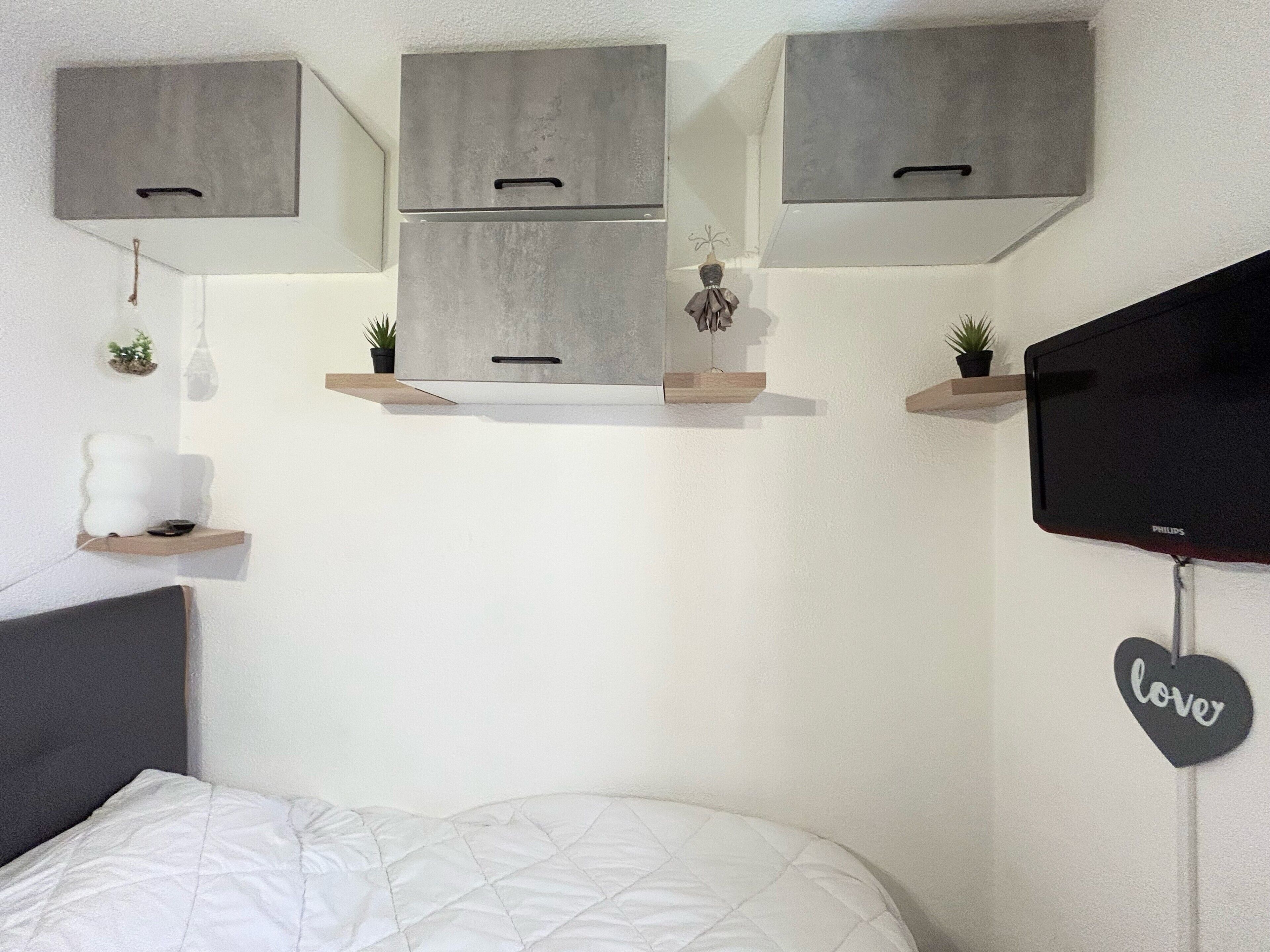 A bedroom with a bed, a wall-mounted TV, and built-in cabinets above a shelf with plants.