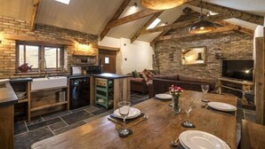 A rustic kitchen with exposed beams, a dining area with a wooden table set for two, and a cozy living space with a fireplace. - Lavender - Barn Conversion - Blossom Farm (Haverfordwest)