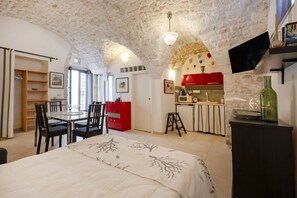 A compact room with a stone ceiling, a dining area with chairs and a table, a bed with a patterned bedspread, a kitchenette with red cabinets, and a small stool.