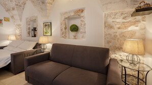 Ein gemütliches Zimmer mit braunem Sofa, einem Bett und einem kleinen Tisch mit Lampe. An den Steinwänden befindet sich eine dekorative Pflanze.