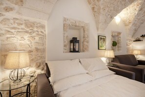 Un dormitorio con pared de piedra, una cama con ropa de cama blanca, dos mesitas de noche con lámparas y una ventana pequeña.