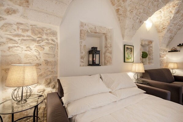 A bedroom with a stone wall, a bed with white linens, two bedside tables with lamps, and a small window.