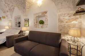 A cozy room with a brown sofa, a bed, and a small table with a lamp. There are stone walls and a decorative plant on the wall.