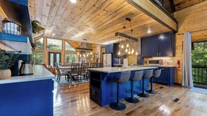 A spacious kitchen with a central island, bar stools, and a dining area with chairs. - Cozy Luxe Cabin Hot Tub+Firepit (Broken Bow)