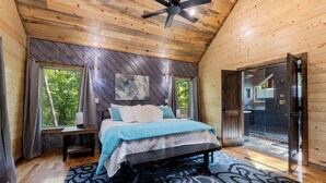 A bedroom with a bed, a nightstand, a window, and a door leading to a bathroom. - Cozy Luxe Cabin Hot Tub+Firepit (Broken Bow)