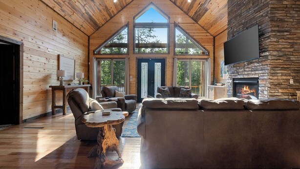 A spacious living room with a stone fireplace, wooden ceiling, and large windows. - Cozy Luxe Cabin Hot Tub+Firepit (Broken Bow)