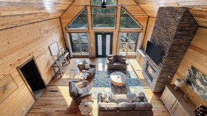 A spacious loft-style room with a high ceiling, large windows, a stone fireplace, and a cozy seating area. - Cozy Luxe Cabin Hot Tub+Firepit (Broken Bow)