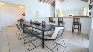 Un comedor con una mesa de madera oscura y sillas de malla metálica, una cocina con armarios blancos y un piso de baldosas.