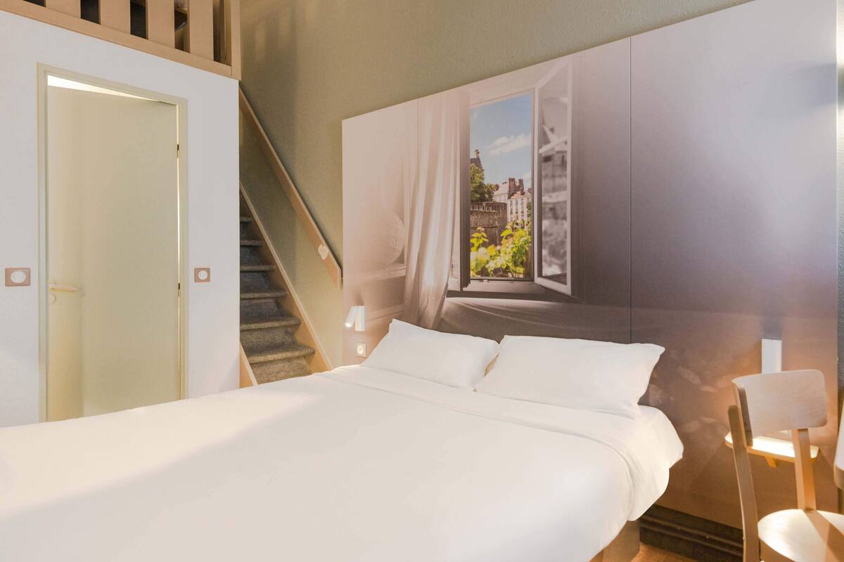 A bedroom with a bed, a window with curtains, a staircase, and a wall-mounted light fixture.