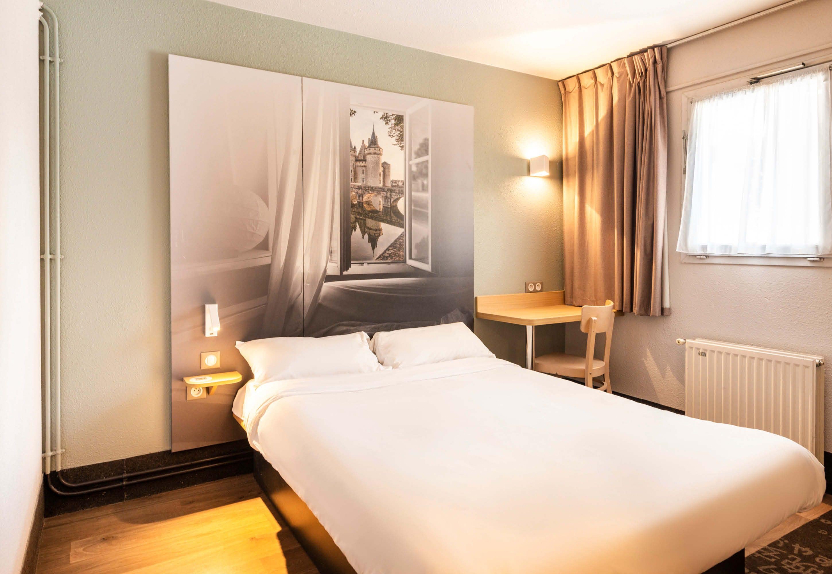 A hotel room with a bed, a nightstand, a chair, a window with curtains, and a wall-mounted light.