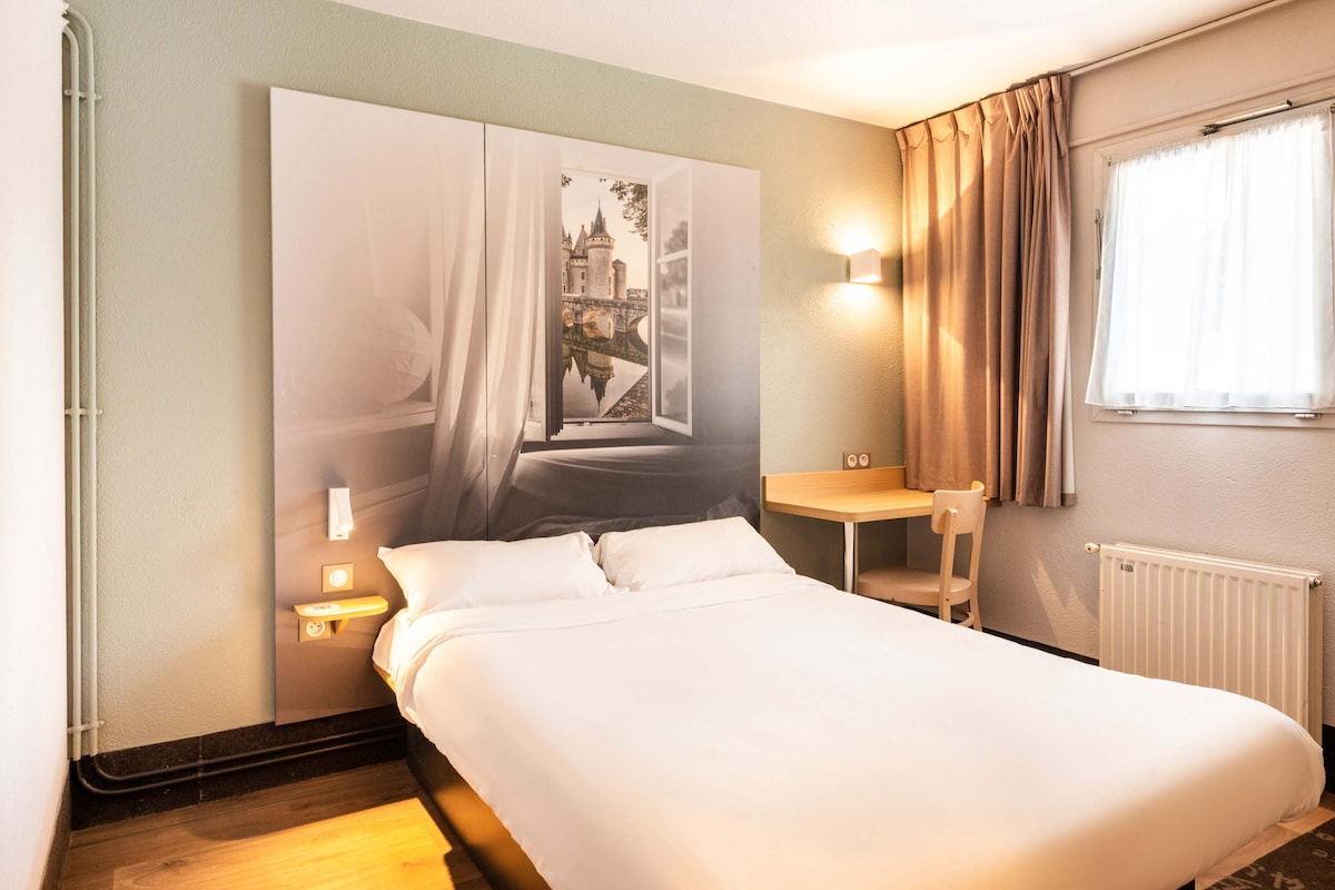 A hotel room with a bed, a nightstand, a chair, a window with curtains, and a wall-mounted light.