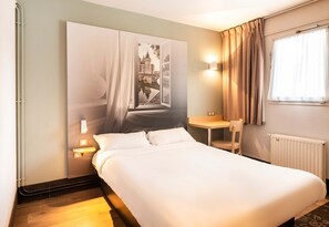 Ein Hotelzimmer mit einem Bett, einem Nachttisch, einem Sessel, einem Fenster mit Vorhängen und einer Wandleuchte.