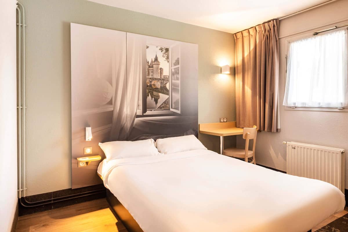 A hotel room with a bed, a nightstand, a chair, a window with curtains, and a wall-mounted light.