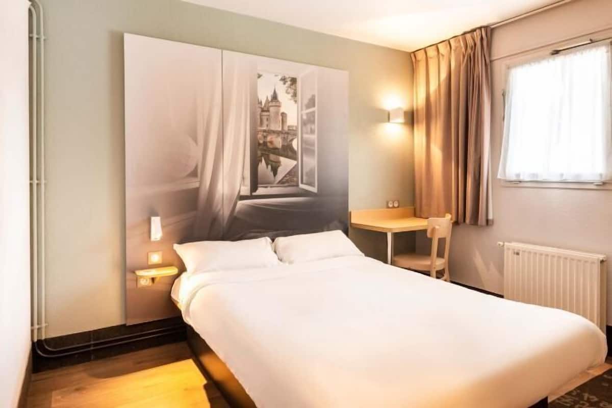 A hotel room with a bed, a desk, a chair, a window with curtains, and a wall-mounted artwork.
