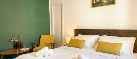 A hotel room with a bed, a painting on the wall, a chair, a small table, and a bedside lamp.