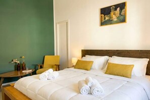 A hotel room with a bed, a painting on the wall, a chair, a small table, and a bedside lamp.